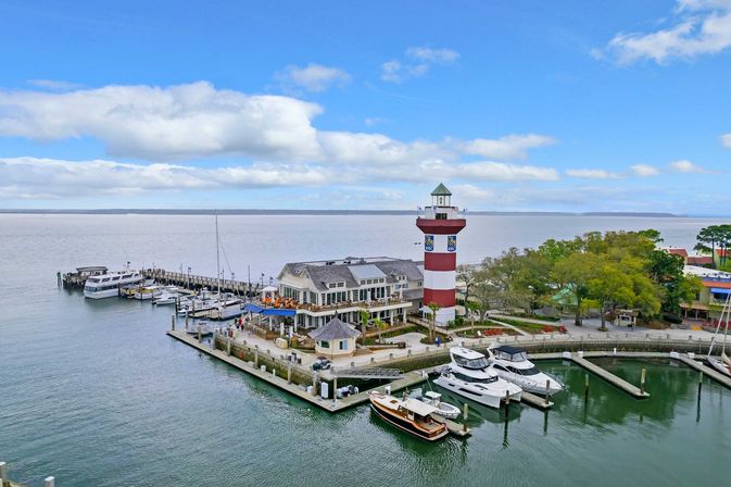 Aerial view of a coastal marina with yachts docked beside waterfront restaurants and a charming red-and-white striped lighthouse overlooking calm harbor waters under a blue sky.