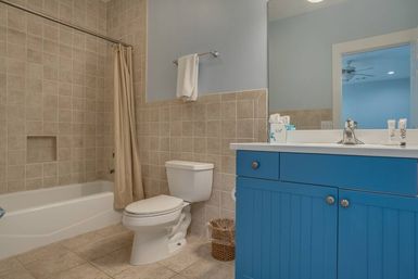 Cheerful bathroom with beige tiled tub-shower and tan curtain, white toilet, wicker wastebasket, and vibrant blue vanity with sink and mirror