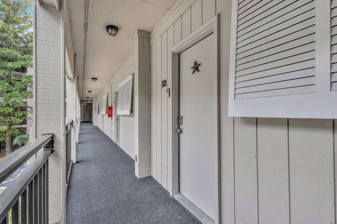 Exterior second-floor balcony corridor in a residential complex with white paneled doors (one with a star), carpeted walkway, metal railing and trees beyond.