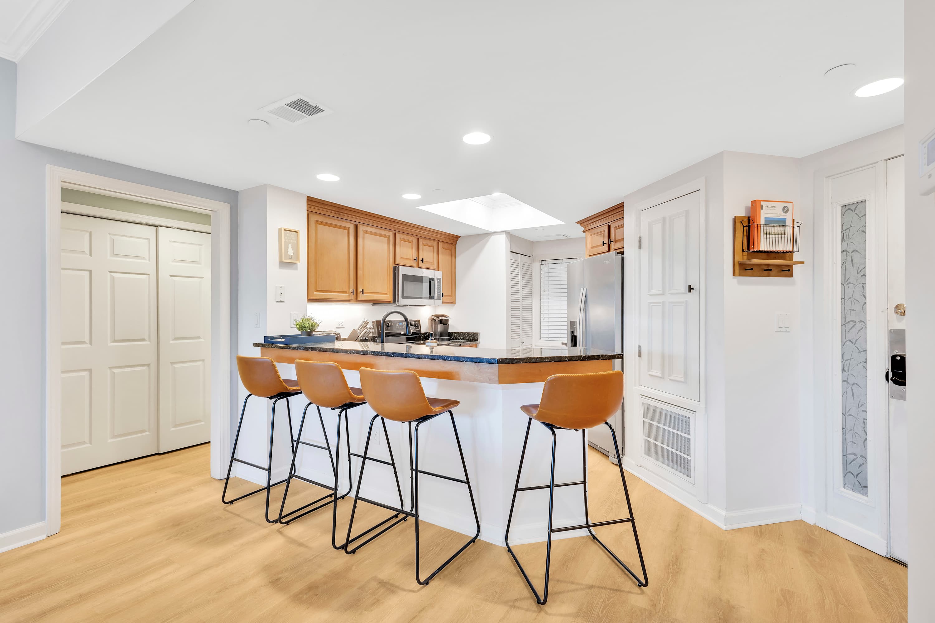 Bright open-concept kitchen with granite breakfast bar and four tan leather bar stools, maple cabinets, stainless appliances, and light wood flooring.
