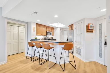 Bright open-concept kitchen with granite breakfast bar and four tan leather bar stools, maple cabinets, stainless appliances, and light wood flooring.