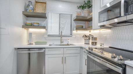 Bright modern white kitchen with stainless steel appliances, hex-tile backsplash, floating wood shelves, and a sink under a window with blinds.