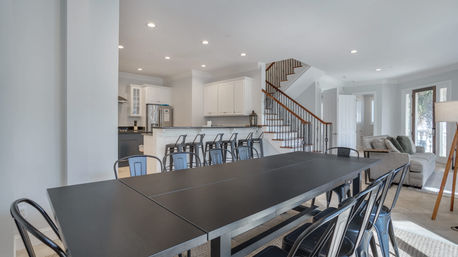 Bright open-concept modern home interior: long black dining table with metal chairs, white kitchen with breakfast bar and stools, staircase and cozy living area with sofa by sliding doors.