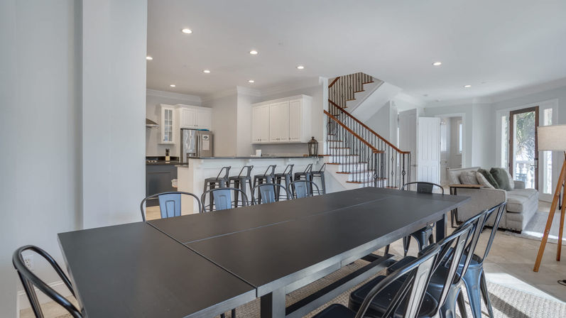 Bright open-concept modern home interior: long black dining table with metal chairs, white kitchen with breakfast bar and stools, staircase and cozy living area with sofa by sliding doors.