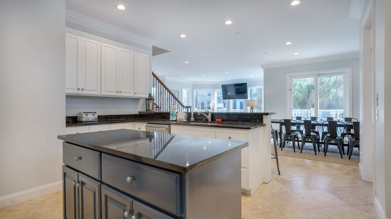 Bright open-plan kitchen with dark granite island and countertops, white cabinets, breakfast bar, stainless sink, wall-mounted TV, adjacent dining table with black metal chairs and sliding glass doors with palm-tree views.
