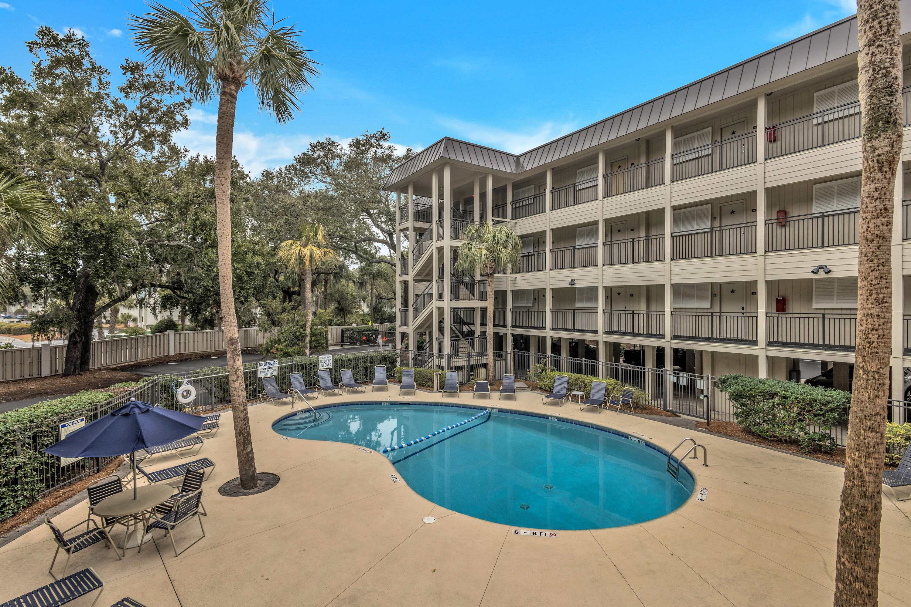 Kidney-shaped outdoor hotel pool in a palm-lined courtyard with lounge chairs, patio tables and umbrellas, and a four-story balcony hotel building under a blue sky