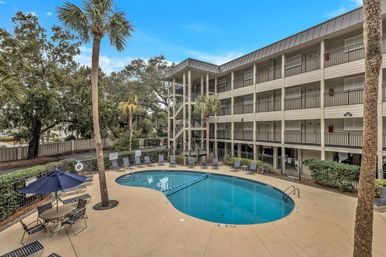 Kidney-shaped outdoor hotel pool in a palm-lined courtyard with lounge chairs, patio tables and umbrellas, and a four-story balcony hotel building under a blue sky