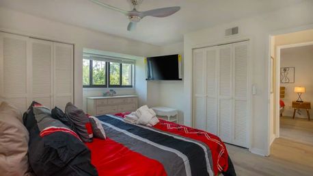 Bright modern bedroom interior with bold red, black, and gray striped bedding on a cozy bed, ceiling fan, wall-mounted TV, white louvered closets, dresser under a sunlit window, and an open doorway to an adjoining room.