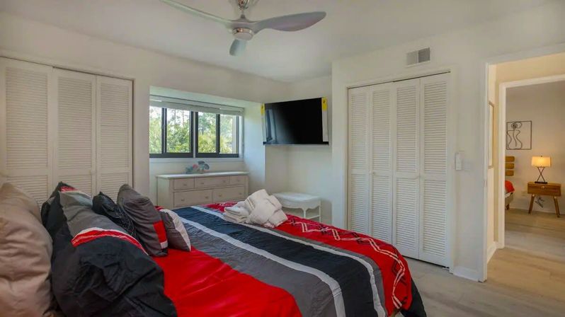 Bright modern bedroom interior with bold red, black, and gray striped bedding on a cozy bed, ceiling fan, wall-mounted TV, white louvered closets, dresser under a sunlit window, and an open doorway to an adjoining room.