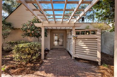 Sunlit brick walkway under a white pergola leading to glass double doors of a beige cottage, flanked by flowering shrubs, a louvered privacy screen with a colorful butterfly ornament, and pine‑straw landscaping.