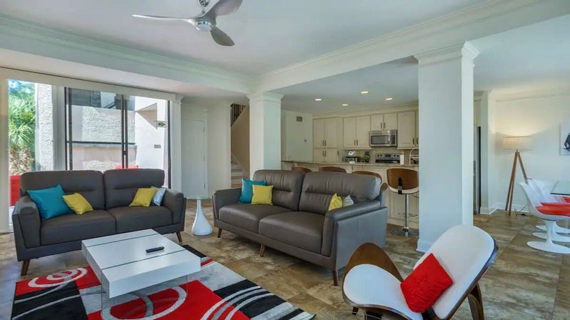 Modern open-plan condo living room and kitchen with gray leather sofas, colorful accent pillows, red-black area rug, white coffee table, bar stools, ceiling fan and sliding glass doors