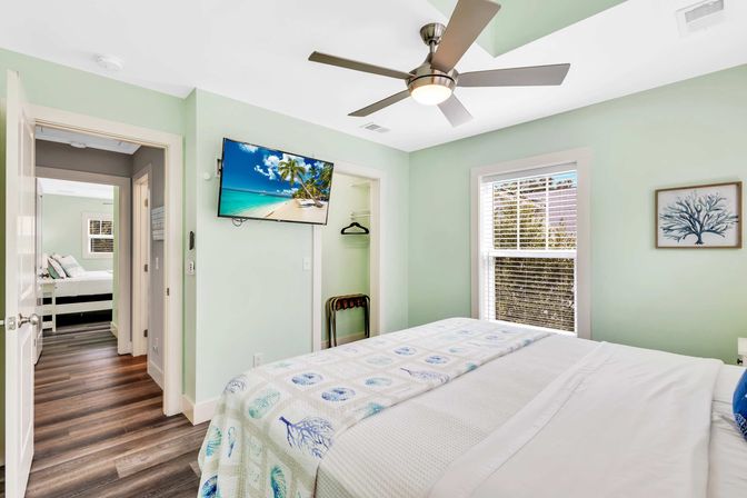 Bright coastal-themed bedroom in a vacation rental with mint-green walls, ceiling fan, wall-mounted TV showing a tropical beach, queen bed with seashell quilt, hardwood floors, sunlit window and open doorway to an adjacent room.