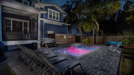 Night backyard of a modern blue two-story house with a glowing purple LED rectangular pool, paver patio with lounge chairs, outdoor sofa, palm trees and wooden fence.