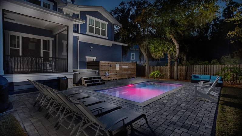 Night backyard of a modern blue two-story house with a glowing purple LED rectangular pool, paver patio with lounge chairs, outdoor sofa, palm trees and wooden fence.