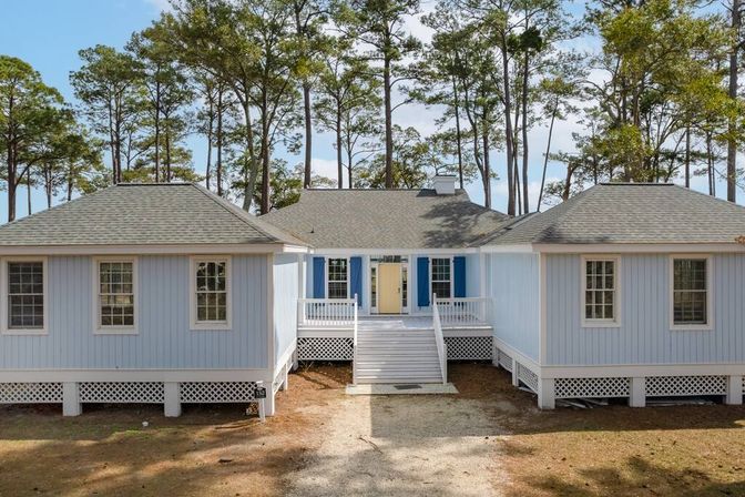 Cheerful light-blue raised coastal cottage with a yellow front door and blue shutters, central staircase up to a white porch, set among tall pine trees with a sandy driveway.