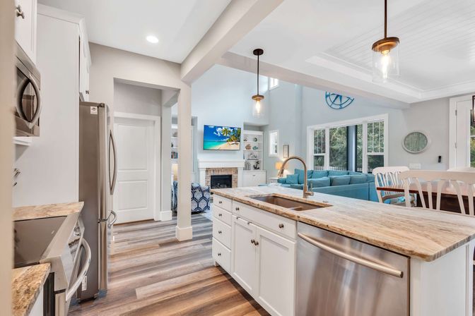 Bright open-concept coastal-inspired kitchen with large marble-look island and sink, stainless steel appliances, wood-plank floors, pendant lights, and adjoining living room with teal sofa, fireplace and large windows