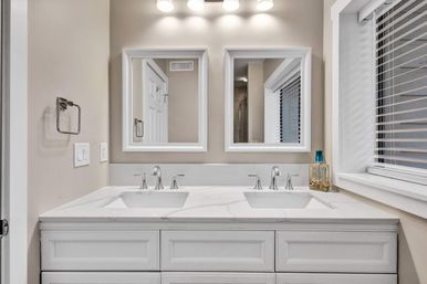 Bright modern residential bathroom with a white double-sink vanity, marble countertop, chrome faucets, dual framed mirrors, overhead vanity lights, and a window with blinds.