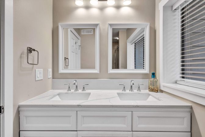 Bright modern residential bathroom with a white double-sink vanity, marble countertop, chrome faucets, dual framed mirrors, overhead vanity lights, and a window with blinds.
