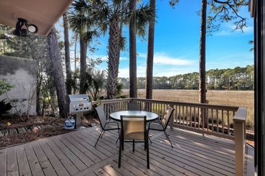 Wooden deck patio with round glass table and four mesh chairs, stainless gas grill beside palm and pine trees overlooking a golden coastal marsh and treeline under a bright blue sky