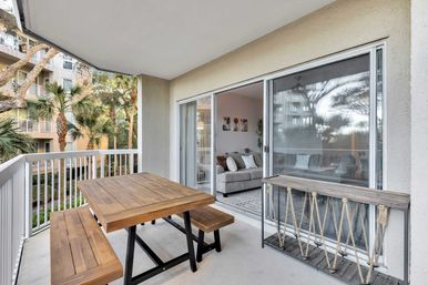 Covered apartment balcony with wooden picnic-style table and benches, sliding glass doors to a cozy living room, and palm trees visible outside for a relaxed, tropical vibe.
