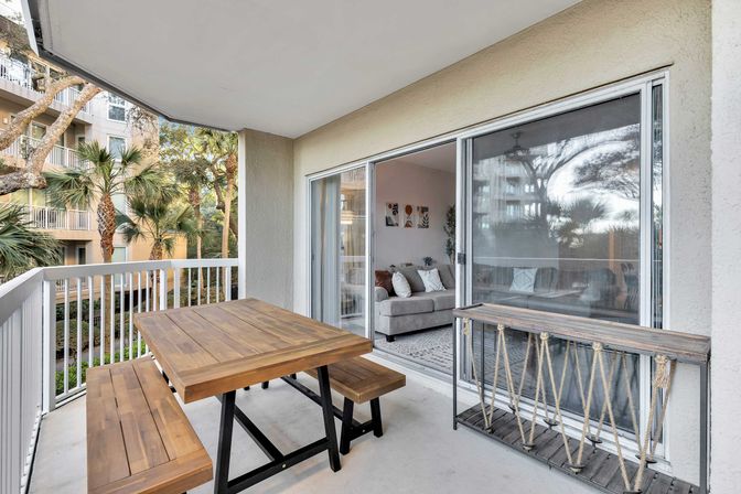 Covered apartment balcony with wooden picnic-style table and benches, sliding glass doors to a cozy living room, and palm trees visible outside for a relaxed, tropical vibe.