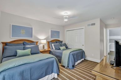 Bright guest bedroom with two double beds in blue quilts and green throws, wood headboards, nightstand lamp, ceiling fan, striped rug and closet