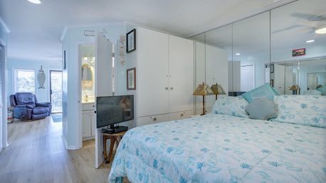 Bright beach-inspired master bedroom with blue coral-print bedding, mirrored closet doors and white built-in cabinets, small TV on a wicker stand, bedside lamps, and an open doorway leading to a seating area with a recliner and balcony windows.
