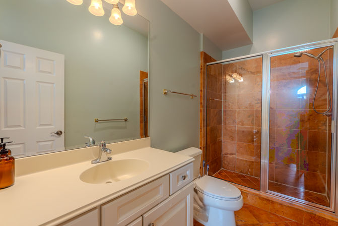 Bright residential bathroom with white vanity and chrome faucet, large mirror, glass-enclosed brown stone-tile shower and toilet