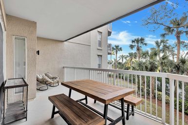 Covered beach condo balcony with wooden picnic table and benches, cozy rattan lounge chairs, white railing and a view of palm trees under a bright blue sky.