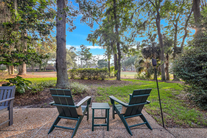 Backyard patio with two green Adirondack chairs and a small table under oak trees draped in Spanish moss, overlooking a sunlit golf fairway and lush landscaping.