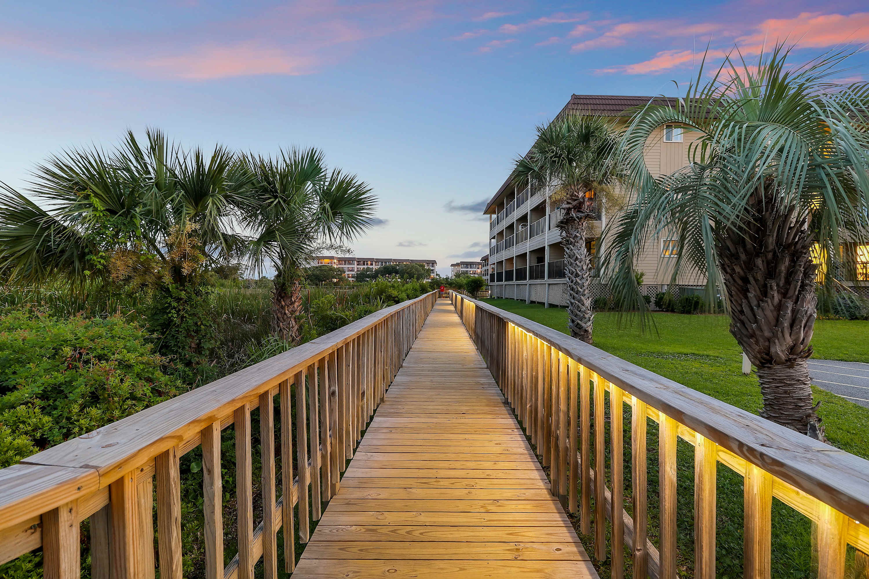 Inviting wooden coastal boardwalk at sunset with warm railing lights, palm trees, and beachside condos in view