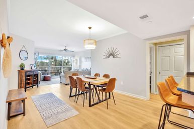 Sunlit open-plan coastal condo with wood floors, dining table and six brown chairs, leather bar stools at the counter, living area and sliding glass doors to a balcony overlooking palm trees.