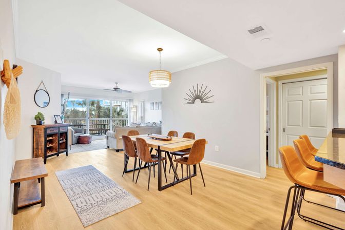 Sunlit open-plan coastal condo with wood floors, dining table and six brown chairs, leather bar stools at the counter, living area and sliding glass doors to a balcony overlooking palm trees.