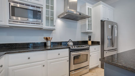 Bright modern kitchen with white cabinets, stainless steel microwave, range, and French-door refrigerator, black granite countertops, stainless range hood, island, kettle and neatly arranged utensils.