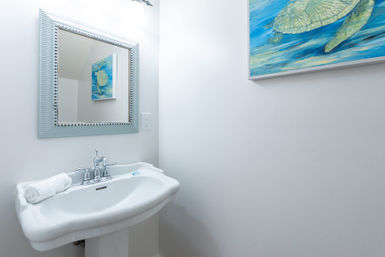 Bright coastal powder room with a white pedestal sink, chrome faucet, rolled towel, beaded framed mirror and sea turtle wall art.
