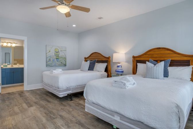 Bright coastal-inspired guest bedroom with two queen beds, wooden headboards, crisp white linens and blue accent pillows, bedside lamp and ceiling fan, wood-look floors and an en-suite bathroom with blue vanity