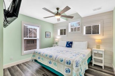 Bright coastal-style beach cottage bedroom with king bed and sea-themed quilt, mint-green walls, shiplap accent wall with three small windows, ceiling fan, wall-mounted TV, white nightstands and wood-look flooring.