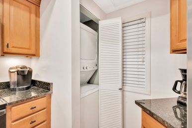 Small modern kitchen with granite countertops and light wood cabinets, showing Keurig and drip coffee makers and a stacked washer-dryer tucked behind a white louvered closet door.