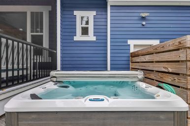 Cozy outdoor hot tub filled with clear water on a small backyard deck against blue house siding, flanked by a wooden privacy fence and black metal railing.