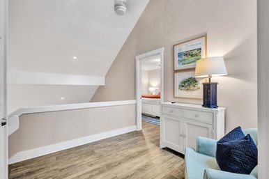 Cozy upstairs landing with sloped ceiling and wood floors, white console table with a navy-base lamp and framed landscape prints, a light-blue armchair with a navy pillow, and an open doorway into a bedroom.