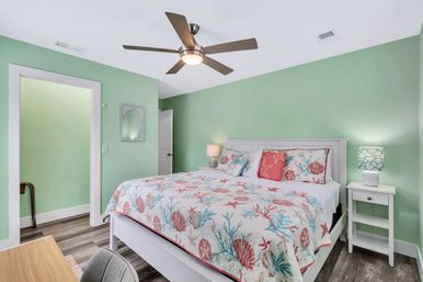 Coastal-themed bedroom with mint-green walls, white bed frame, shell-and-starfish quilt and pillows, modern ceiling fan, bedside lamps and wood-look flooring.