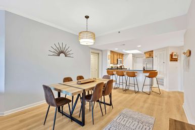 Open-concept dining room and kitchen in a modern apartment — light wood floors, rectangular wood dining table with six brown upholstered chairs, woven pendant light, sunburst wall decor, and a breakfast bar with four tan leather bar stools in front of wood cabinets and a stainless steel fridge.