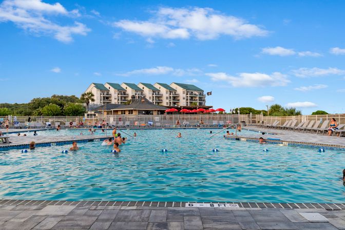 Sunny outdoor resort pool with people swimming, multi-story hotel with green roofs and balconies in the background, lounge chairs and red umbrellas on the deck