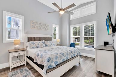 Bright coastal-style bedroom with white wood bed and blue coral bedding, matching nightstands and lamps, ceiling fan, large windows with white blinds, hardwood floors and wall-mounted TV