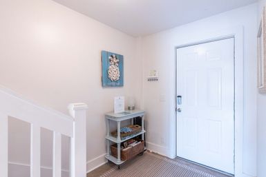 Bright coastal-style entryway with white front door and staircase, small blue two-shelf console holding woven baskets and lamp, striped rug and pineapple "welcome" wall art.