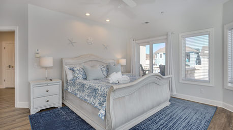 Sunny coastal-style master bedroom in a vacation home with a white sleigh bed, blue floral bedding, starfish wall decor, matching nightstand and blue rug, sliding glass door to a balcony with neighborhood view.
