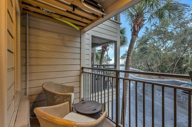 Cozy covered balcony with two wicker chairs and a small round table, surfboards stored overhead, overlooking palm trees, a coastal road and water in the distance.