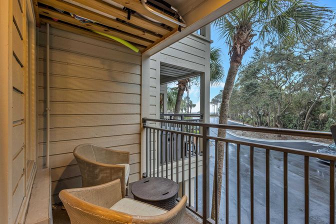 Cozy covered balcony with two wicker chairs and a small round table, surfboards stored overhead, overlooking palm trees, a coastal road and water in the distance.