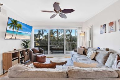 Bright coastal condo living room with a large neutral sectional, leather accent chair and ottoman, round wood coffee table, ceiling fan, wall-mounted TV showing a beach scene, and sliding glass doors opening to a palm-tree lined balcony.