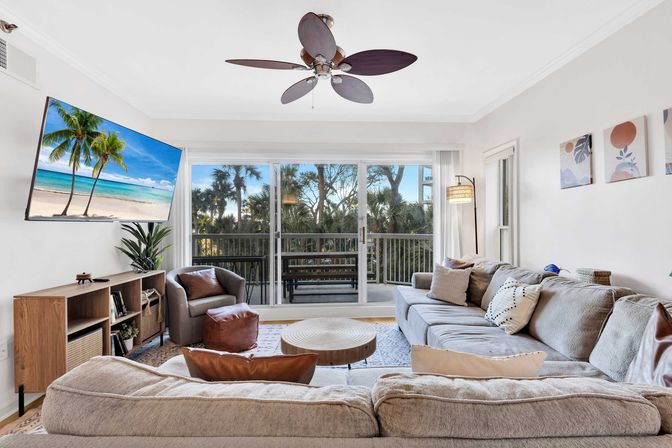 Bright coastal condo living room with a large neutral sectional, leather accent chair and ottoman, round wood coffee table, ceiling fan, wall-mounted TV showing a beach scene, and sliding glass doors opening to a palm-tree lined balcony.